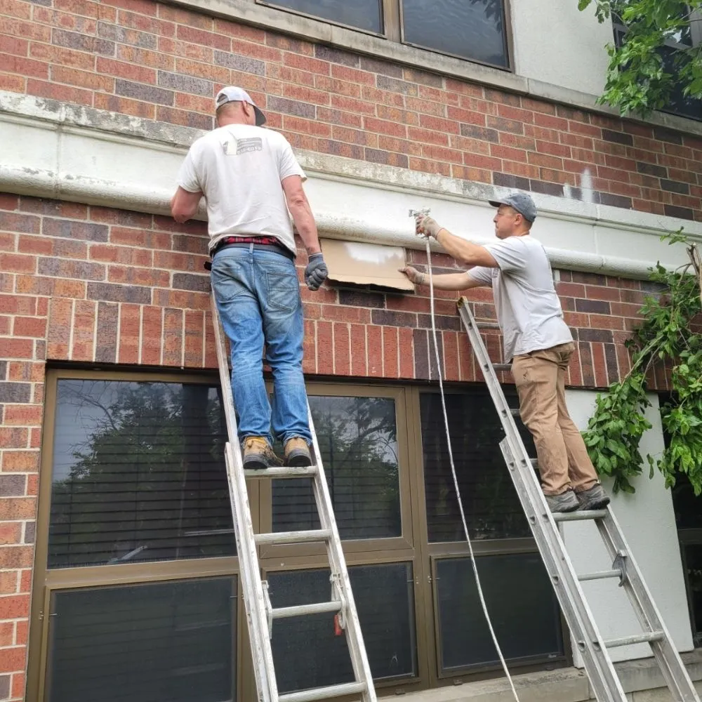 Two workers on ladders tuckpointing brick wall above windows